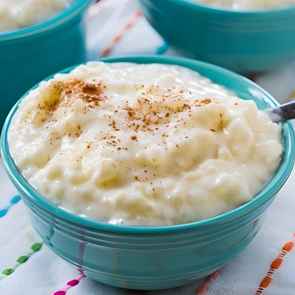 Creamy rice pudding, topped with cinnamon, in a turquoise bowl on a white patterned cloth.
